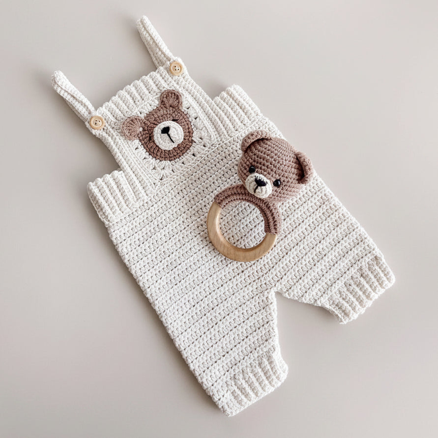 A white crocheted baby romper with a brown bear face applique on the chest and matching brown bear head detail on the shoulder strap, styled with a wooden teething ring toy featuring a brown bear head. The romper is laid flat on a light beige surface with neutral decorative props.