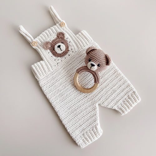 A white crocheted baby romper with a brown bear face applique on the chest and matching brown bear head detail on the shoulder strap, styled with a wooden teething ring toy featuring a brown bear head. The romper is laid flat on a light beige surface with neutral decorative props.