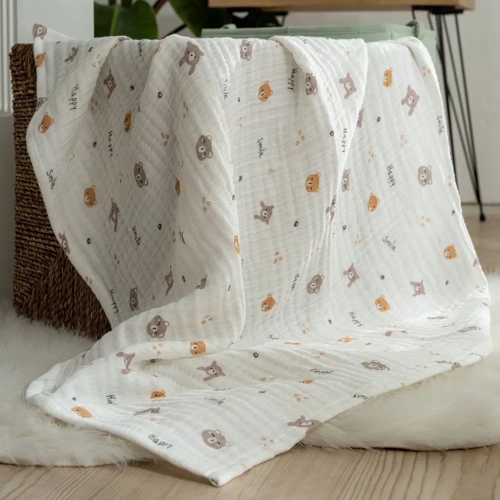 White muslin baby blanket with repeating teddy animal print, spread on cushion and bedding